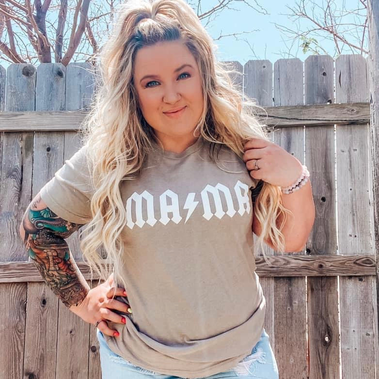 A woman with long, wavy blonde hair stands by a wooden fence outdoors. She’s wearing the MAMA Rocker • Stone Tee with “MAMA” in a rock band–style font and ripped jeans, showing a colorful tattoo sleeve on her right arm.