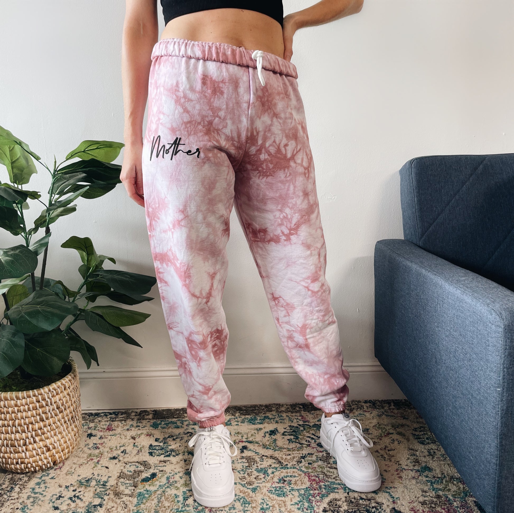 Indoors, someone wears Mother • Rose Dyed Sweats—pink and white tie-dye sweatpants in pre-shrunk cotton with Mother on one leg—paired with white sneakers, standing by a plant and a blue sofa.