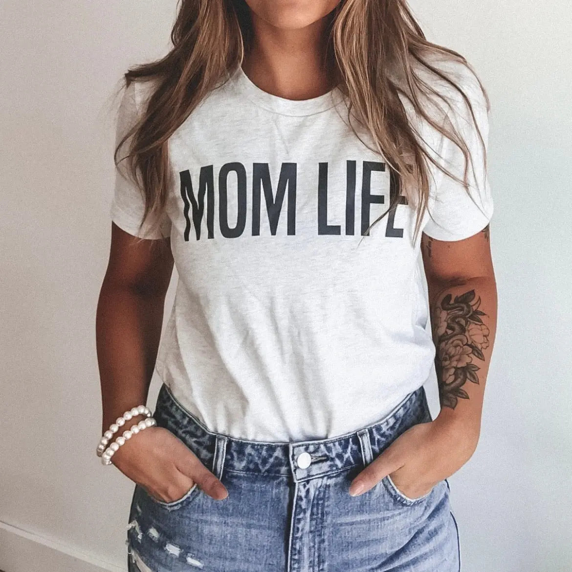 A woman wears the MOM LIFE • Ash Tee with bold lettering, paired with blue high-waisted jeans, a beaded bracelet, and displays a floral tattoo on her left forearm. This unisex Bella Canvas shirt offers both style and comfort. Hands in pockets.