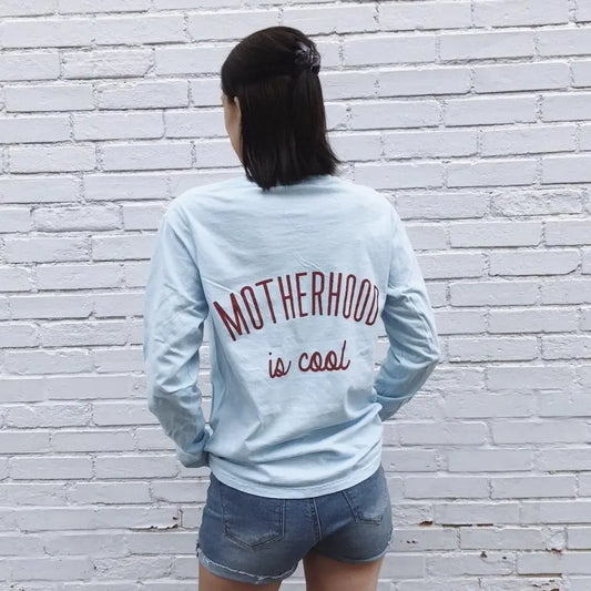 A woman faces a white brick wall, wearing the light blue Motherhood is Cool Back Print • Long Sleeve shirt from the vintage collection with red and black lettering, paired with denim shorts.