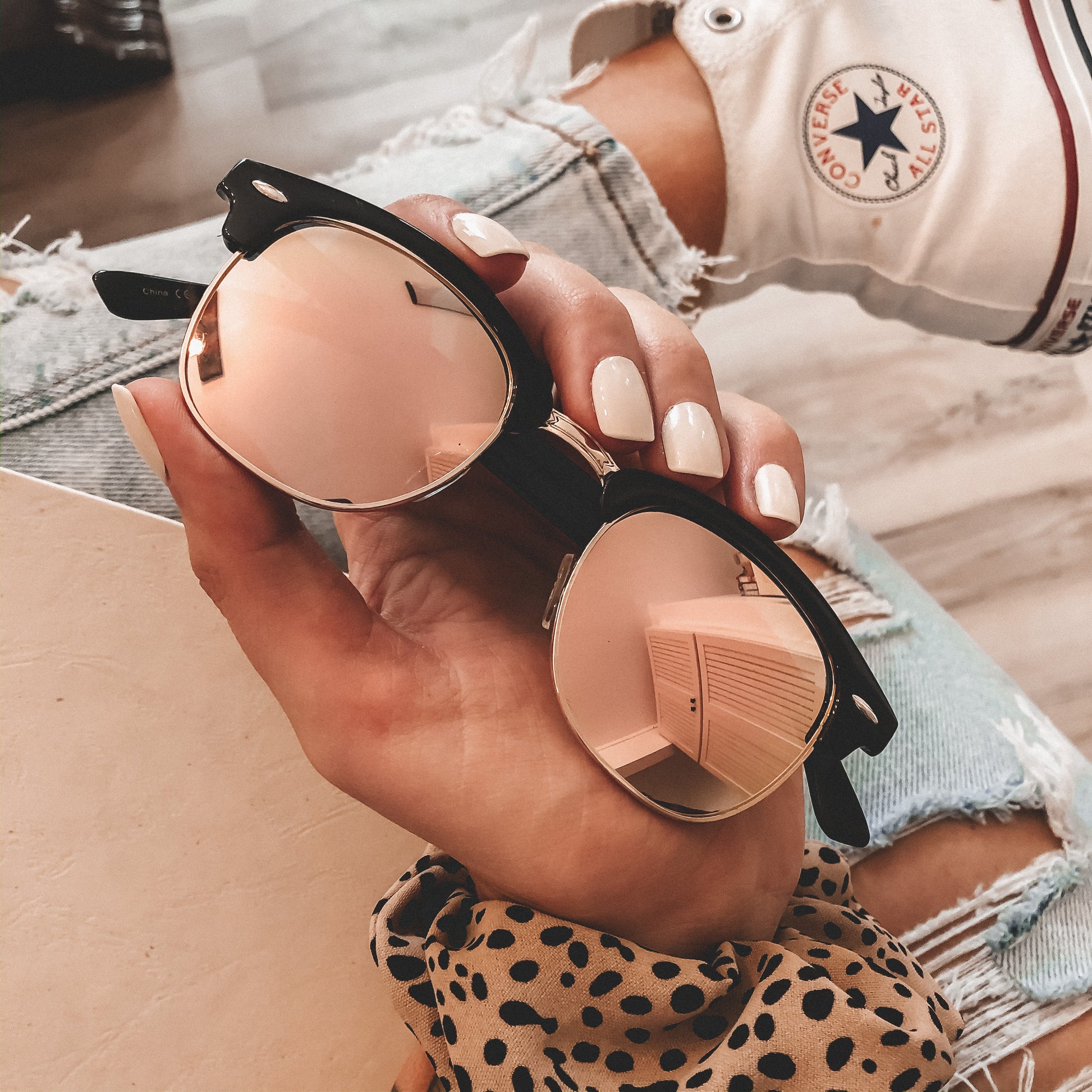 A hand with manicured nails holds Mirror Wayfarer Sunglasses (Adult, More Colors!), featuring UV400 protection and reflective lenses. The person wears ripped jeans, a polka dot top, and white Converse sneakers.