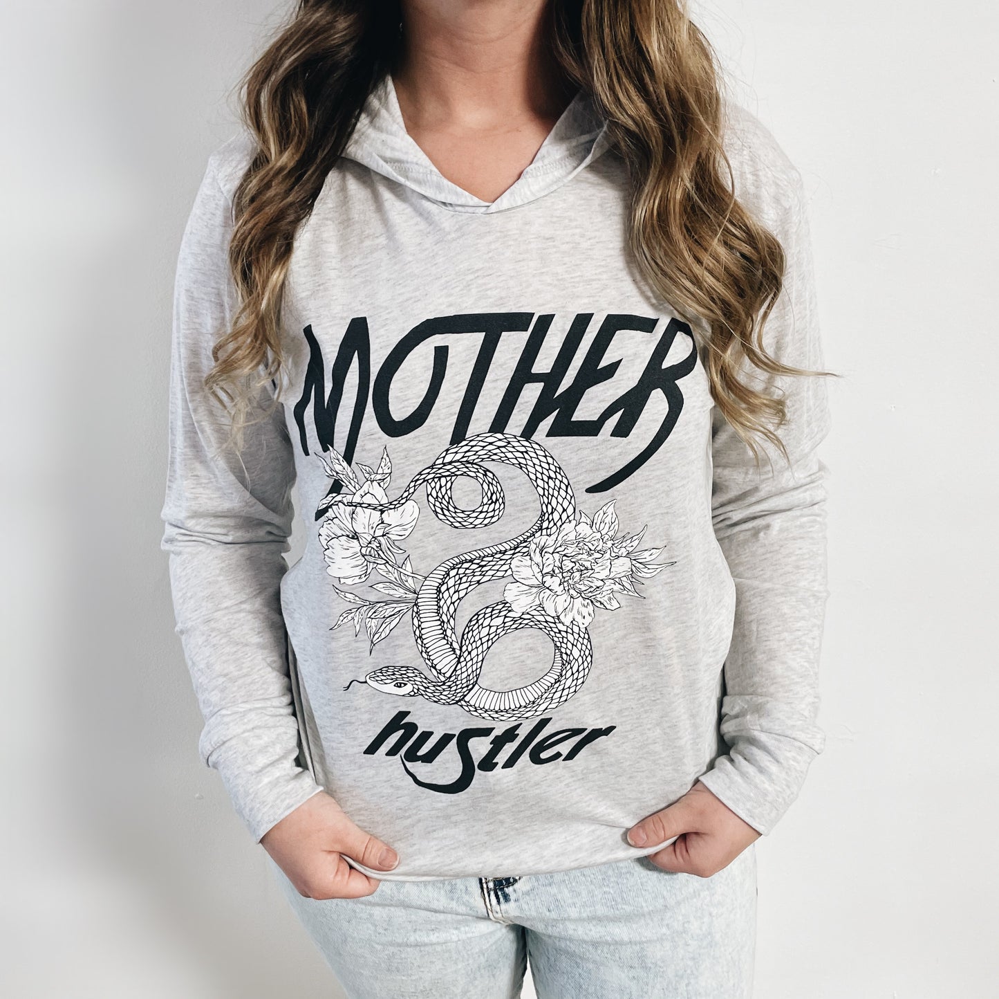 A woman wears the Mother Hustler • Lightweight Hoodie—a lightweight unisex triblend with MOTHER Hustler and a snake among flowers on the front—standing against a plain white background.