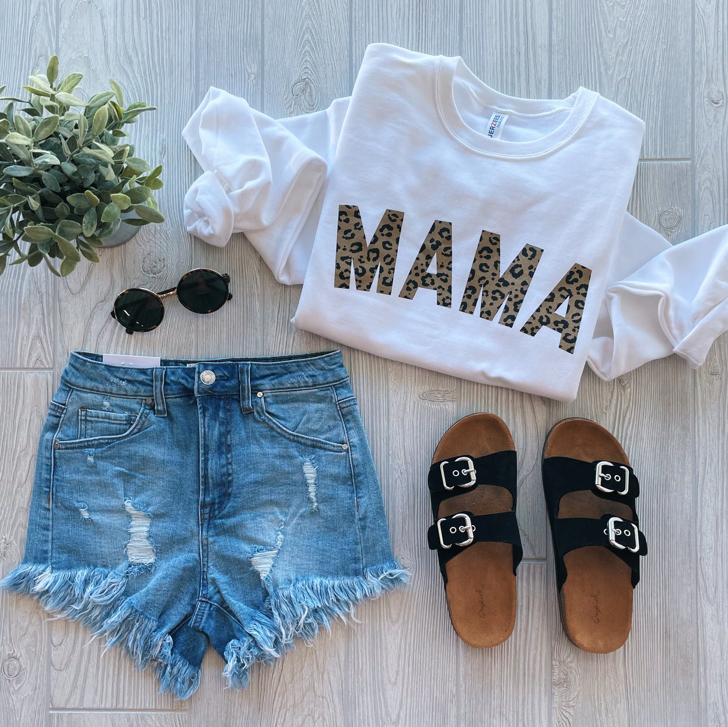 A flat lay of the MAMA Cheetah • White Pullover—comfy unisex fit with leopard print letters—styled with distressed denim shorts, black sandals, round sunglasses, and a green potted plant on a light wood floor.