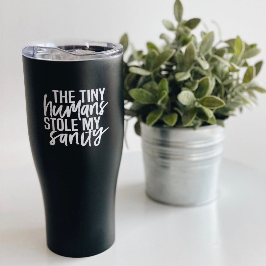 The Tiny Humans • Tumbler, a black stainless steel travel cup printed with The tiny humans stole my sanity, stands beside a potted green plant in a metal pot on a white surface.