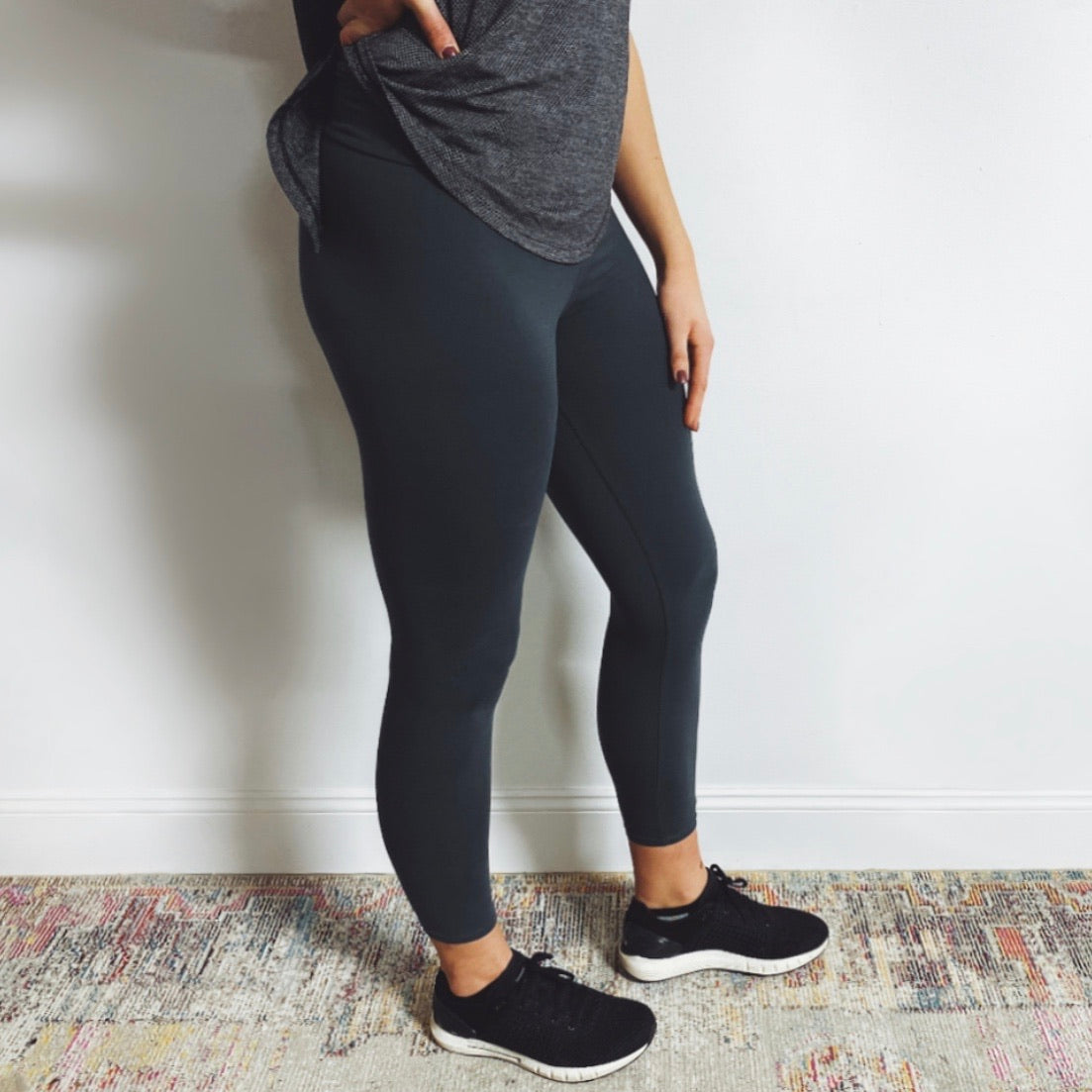 A person, shown from shoulders down indoors against a white wall, wears a gray top and Wide-Waist Leggings • More Colors! in super-soft brushed jersey, paired with black athletic shoes.