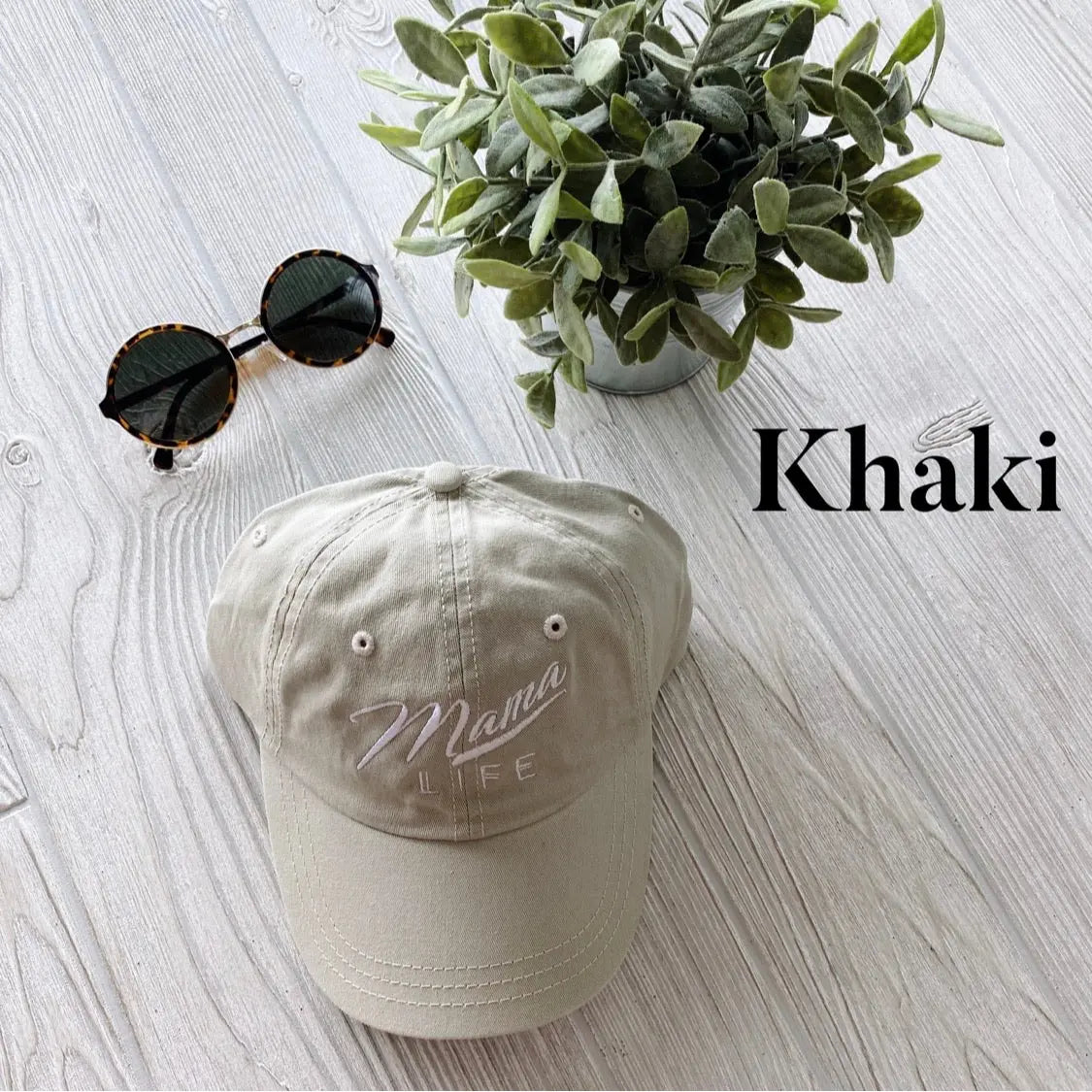 A Mama Life Hat • NEW Colors! in khaki canvas, with vintage Mama Life embroidery on the front, rests on a light wood surface beside round sunglasses and a potted green plant. Khaki is written to the right.