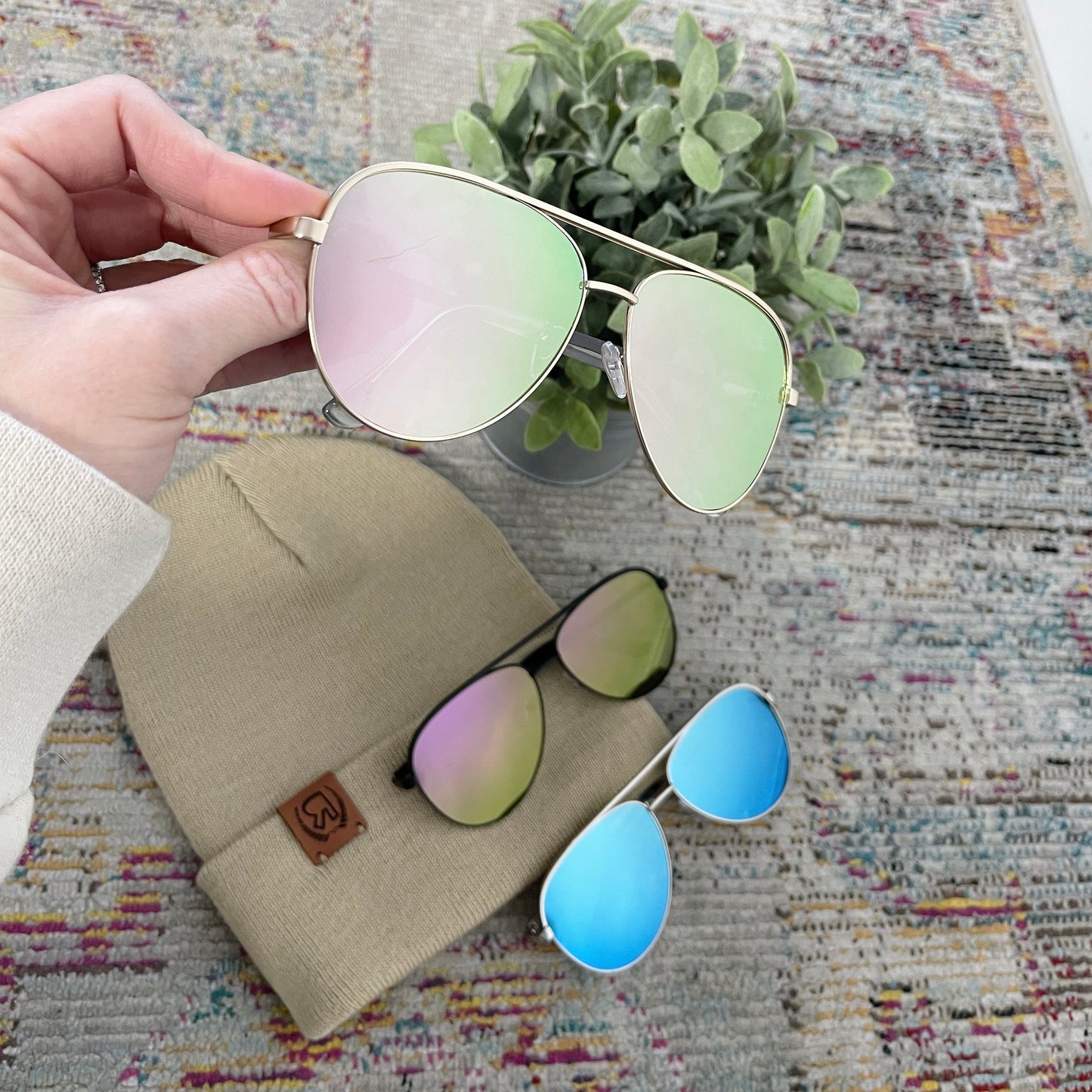 A hand holds gold-framed Bri Sunglasses • Adult with pink-tinted UV400 lenses above a beige beanie on a patterned surface. Nearby are black sunglasses with green lenses and white ones with blue, next to a small plant.