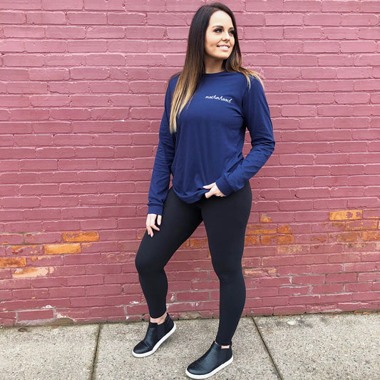 A woman smiles in front of a pink brick wall, looking to the side in the Motherhood Long Sleeve • Navy, paired with black leggings and slip-on shoes—an effortlessly stylish look for moms. One hand rests in her pocket as she enjoys her day.
