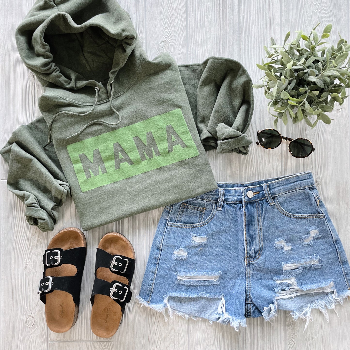 Flat lay of the Mama Box • Olive Tonal: a green fleece sweatshirt hoodie with MAMA suede puff print, paired with distressed denim shorts, black buckle sandals, round sunglasses, and a small potted plant on a light wood surface.