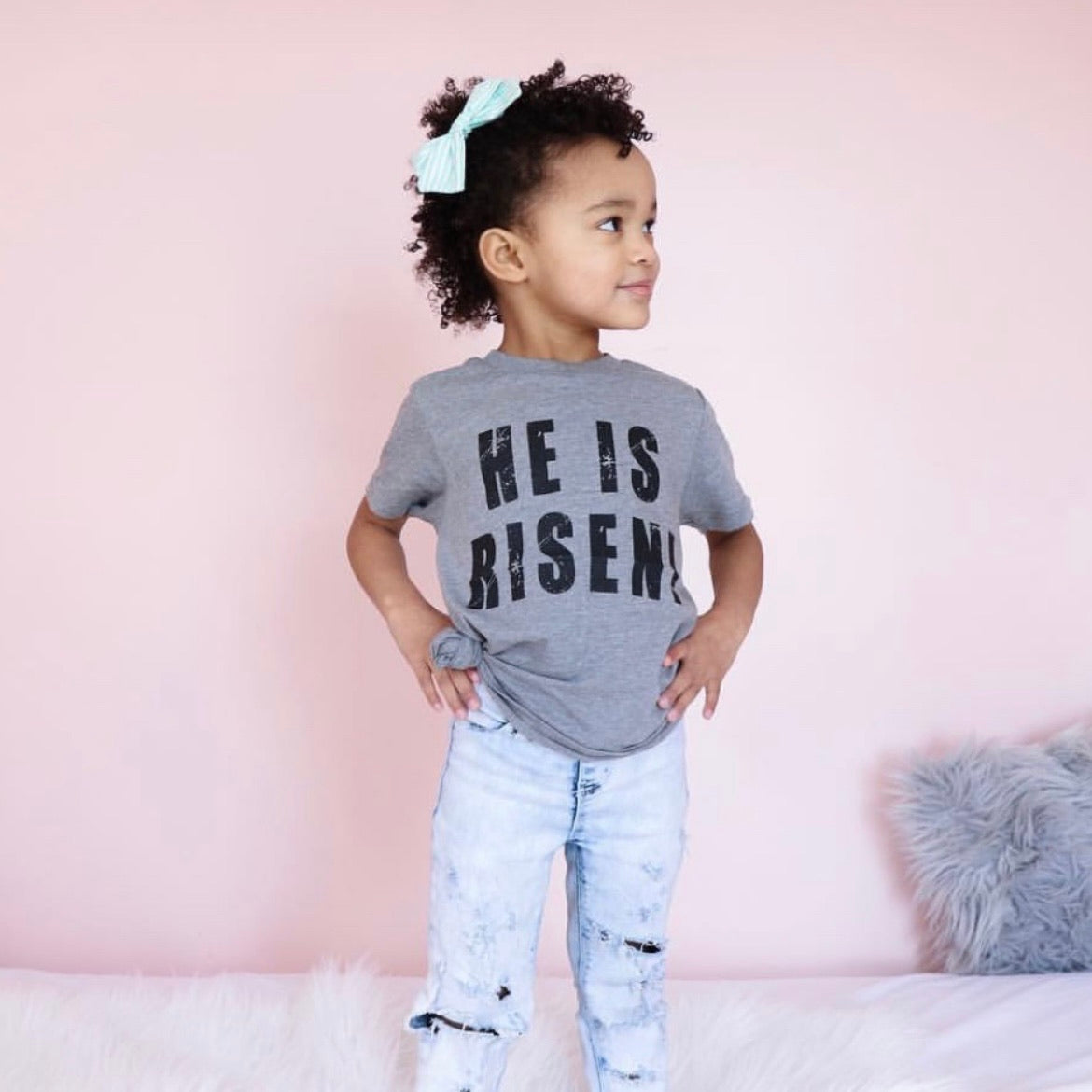 A young child with curly hair and a light blue bow stands on a bed against a pale pink wall, wearing the He is Risen • Kids Tee paired with light blue distressed jeans.
