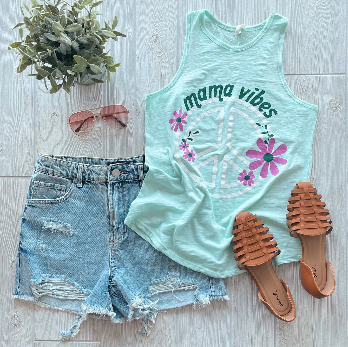 The Mama Vibes • High Neck Tank in light green, featuring a flowy fit and peace sign, styled with distressed denim shorts, brown woven sandals, pink sunglasses, and a small green plant on a light wooden floor.