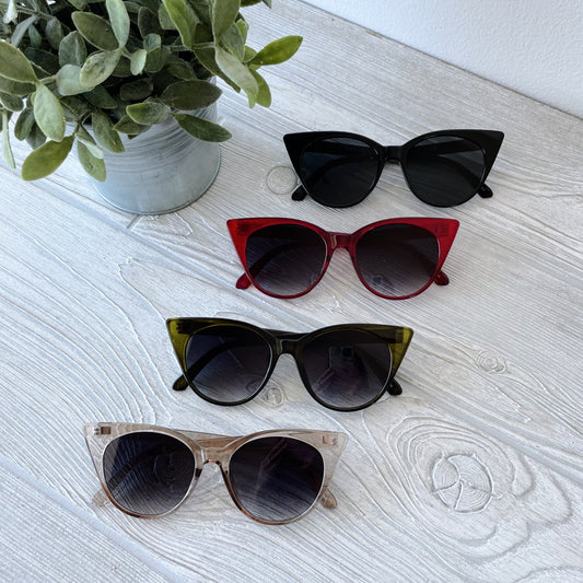 Four pairs of Kat • Adult Sunglasses—black, red, green, and beige cat-eye styles—are arranged vertically on a light wooden surface beside a potted plant. Perfect for elevating any look with bold style.