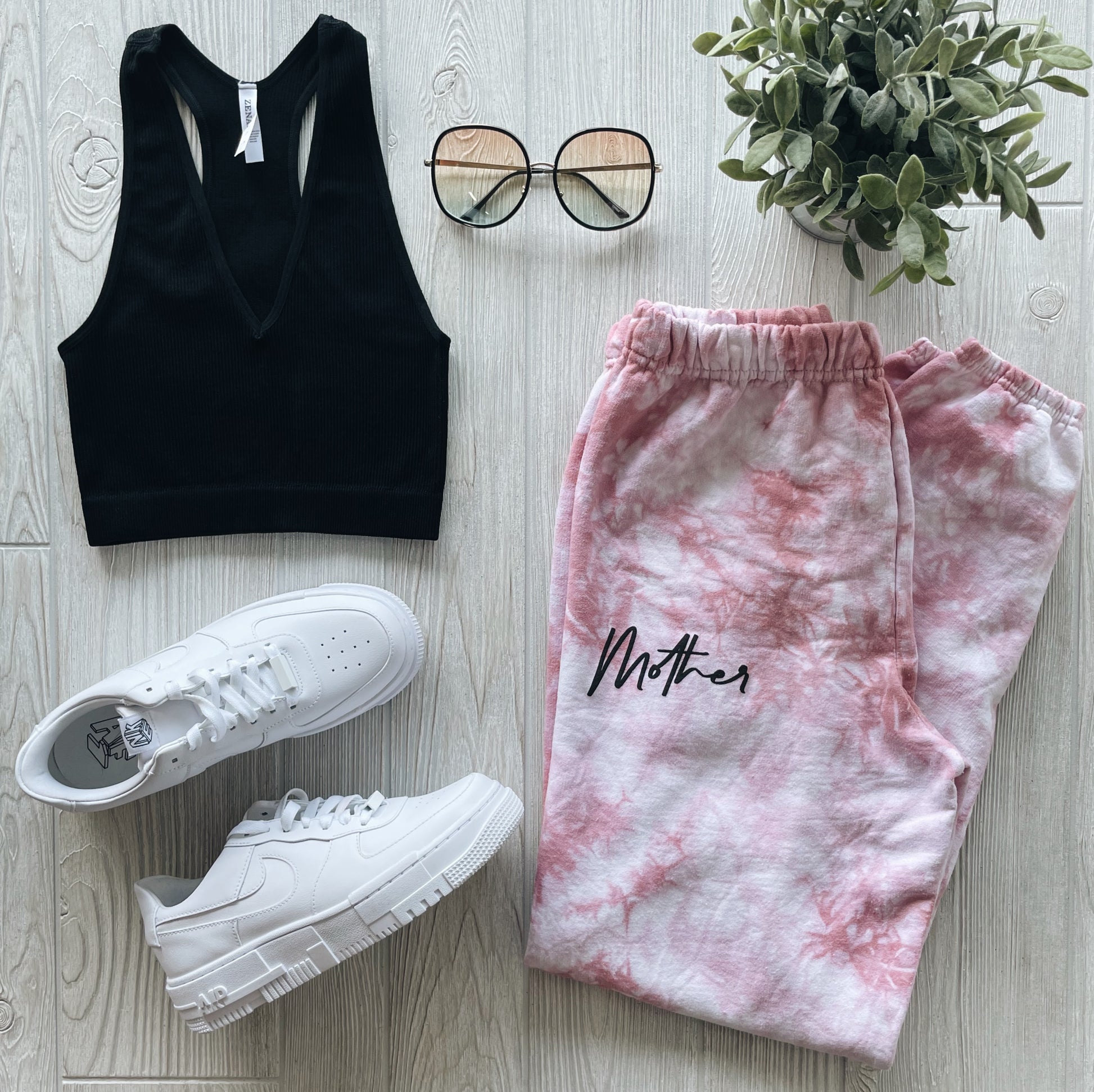 A flat lay of trendy casual wear: a black sleeveless crop top, Mother • Rose Dyed Sweats, white sneakers, round glasses, and a potted green plant on a light wood surface.