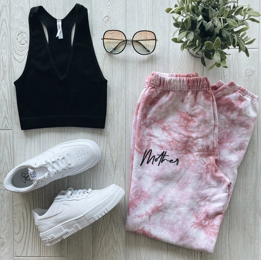 A flat lay of trendy casual wear: a black sleeveless crop top, Mother • Rose Dyed Sweats, white sneakers, round glasses, and a potted green plant on a light wood surface.