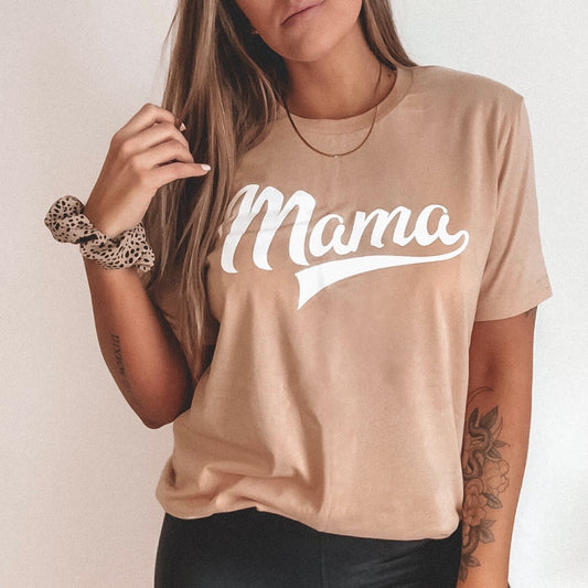 A woman with long brown hair wears the Mama Sporty • Tan Tee—beige with “Mama” in white script—paired with a gold necklace, leopard print scrunchie, black pants, and a floral forearm tattoo.