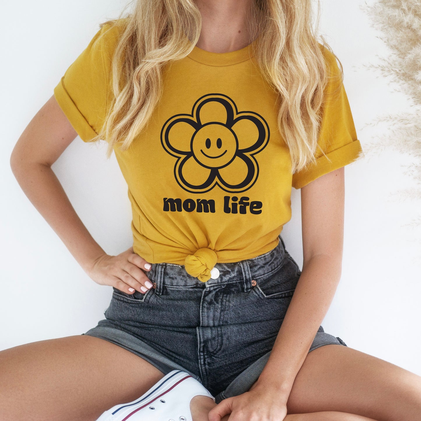 Woman in a Happy Flower • Mustard Tee, featuring a smiling flower graphic, denim shorts, and white sneakers. Made from soft Airlume cotton with a comfy unisex fit. Sitting cross-legged on the floor against a white background.