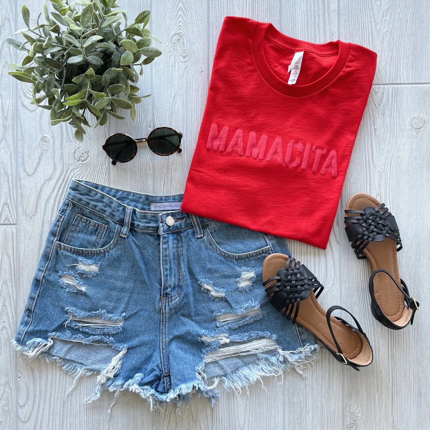 A flat lay of the red MAMACITA • Tonal Poppy Tee in a unisex fit by Bella/Canvas, paired with distressed denim shorts, black woven sandals, round black sunglasses, and a potted green plant on a light wooden surface.