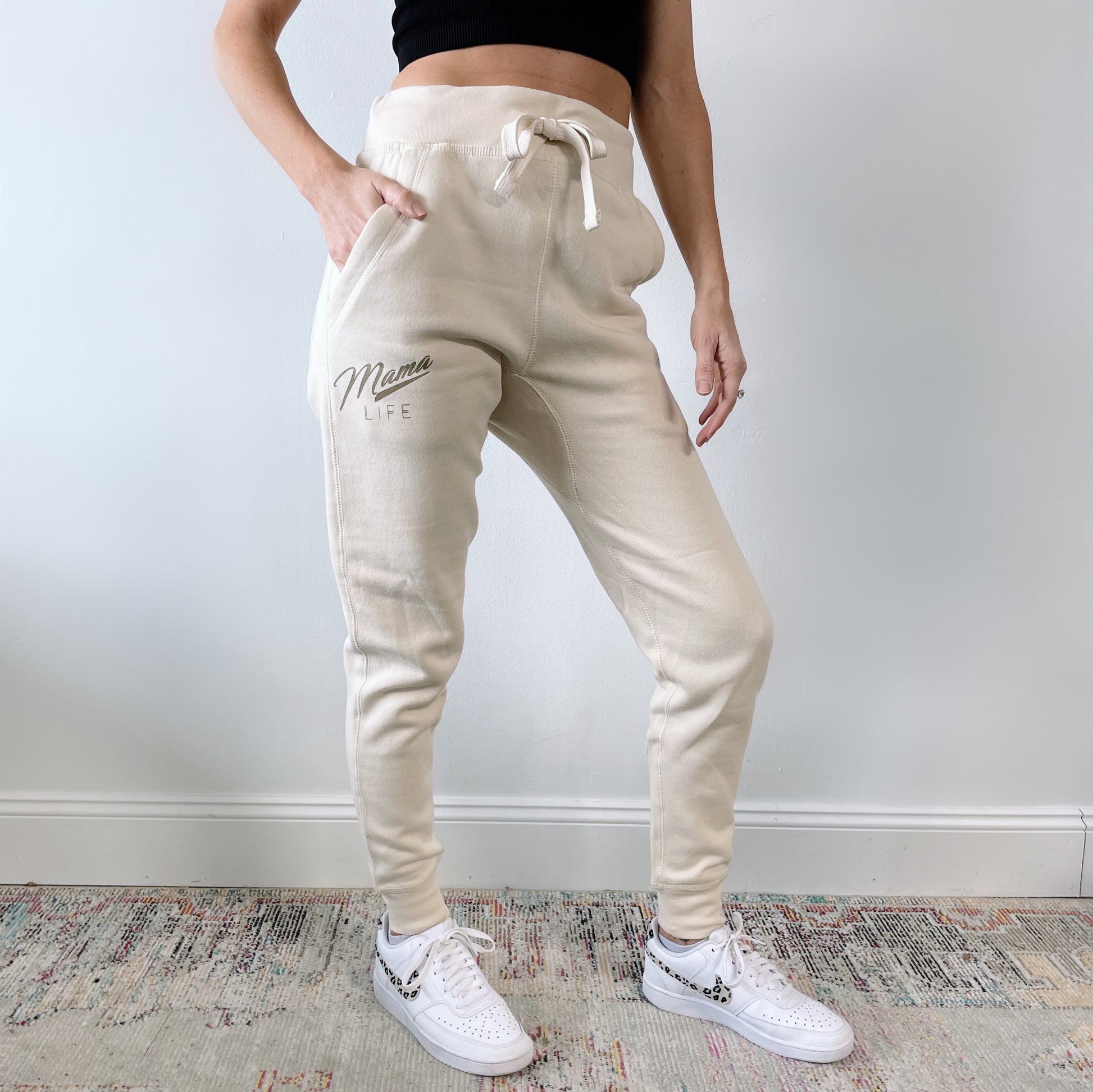 A person wearing Retro Mama Life • Sand Joggers, a black crop top, and white sneakers stands on a patterned rug against a plain light wall.