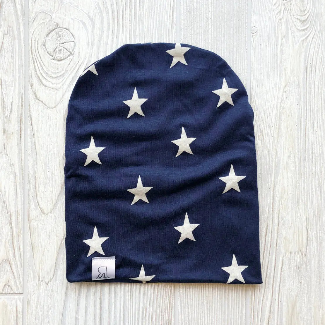 The Star Spangled Beanie is a navy blue handmade hat featuring a pattern of white stars and a small white tag with a black logo at the bottom edge, displayed flat on a light wooden surface.