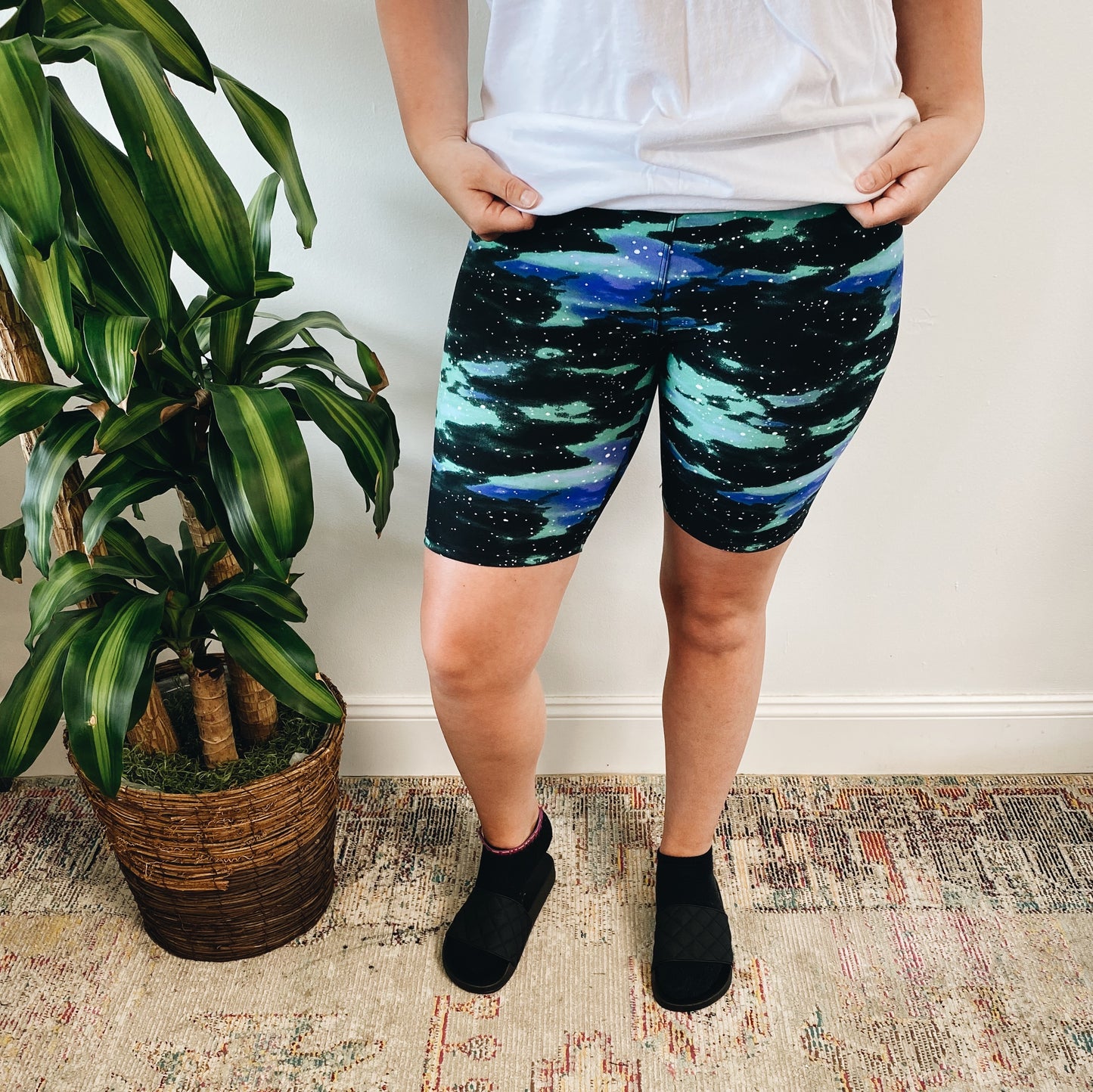 A person stands indoors on a patterned rug, wearing a white t-shirt, Galaxy • Brushed Biker Shorts, and black slippers. A potted green plant sits next to them against a white wall.