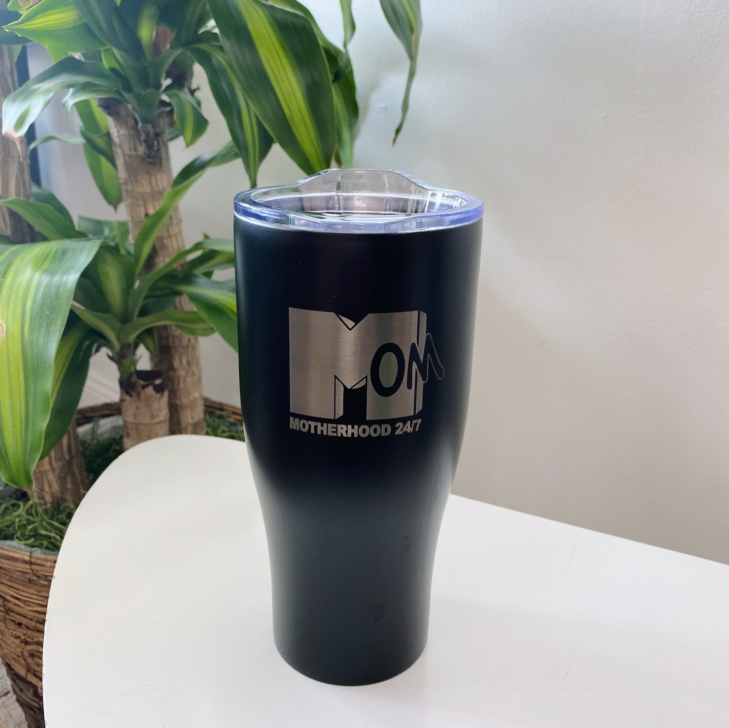 The MTV Mom • Tumbler, a black insulated stainless steel travel mug with a clear lid, is engraved with MOM and MOTHERHOOD 24:7. It sits on a white surface with green leafy plants in the background for a fresh touch.
