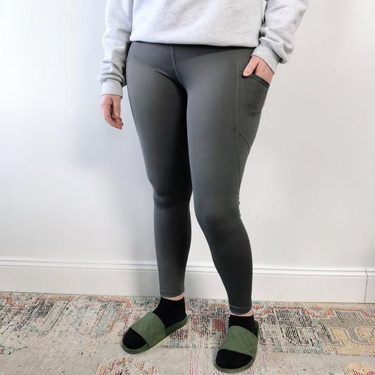 A person stands indoors against a white wall, wearing a light gray sweatshirt and Presley Pocket Legging in dark gray, featuring moisture-wicking fabric and pockets, paired with green open-toe slippers and black socks.