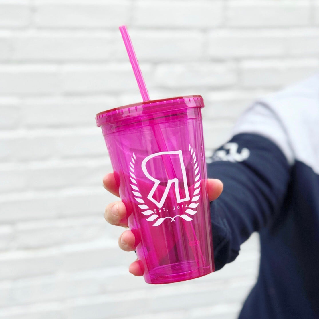 Someone holds the Pink R Logo • Double Wall Tumbler, a transparent pink acrylic cup with double wall insulation and a straw. It features a bold white R logo, laurel leaves, and EST. 2014 on the front, set against a white brick wall background.