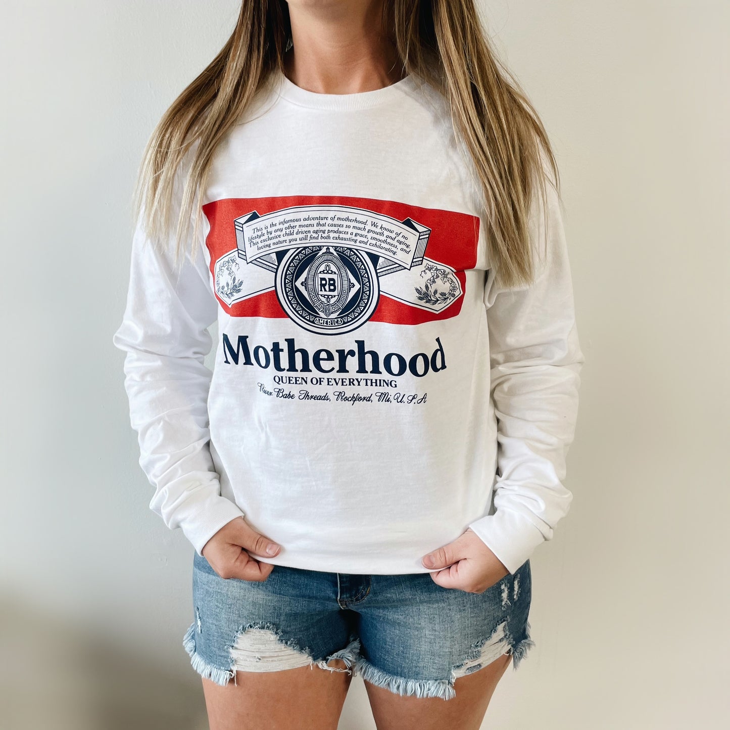 A woman models the Queen of Everything • Long Sleeve, featuring a Budweiser-inspired Motherhood: Queen of Everything graphic, with frayed denim shorts. The unisex sizing offers a great fit for all, shown against a plain white background.