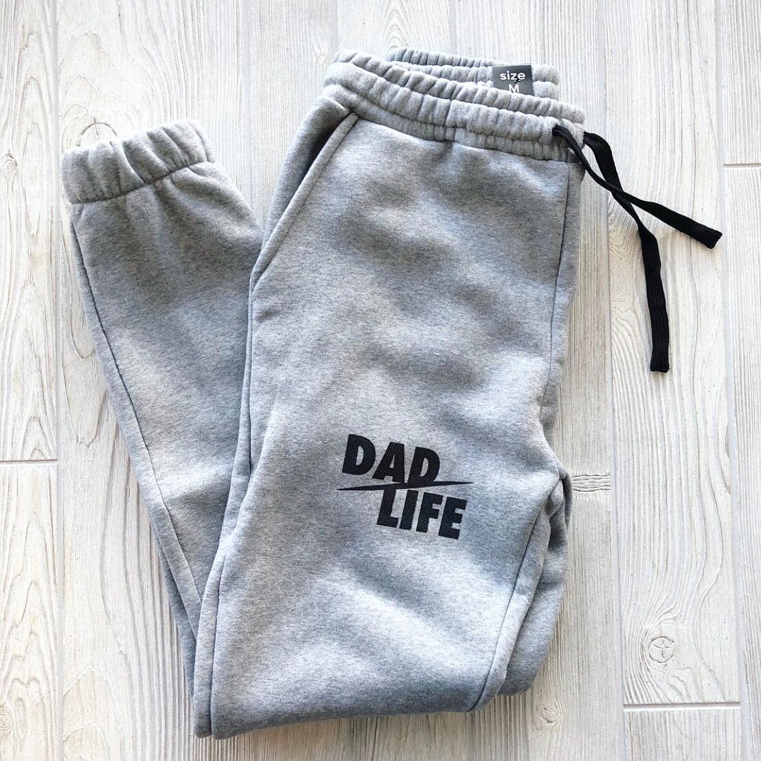 Dad Life Joggers • Gray, shown laid flat on a light wood floor, feature a black drawstring and bold DAD LIFE text on the left leg—perfect cozy loungewear for relaxing at home.