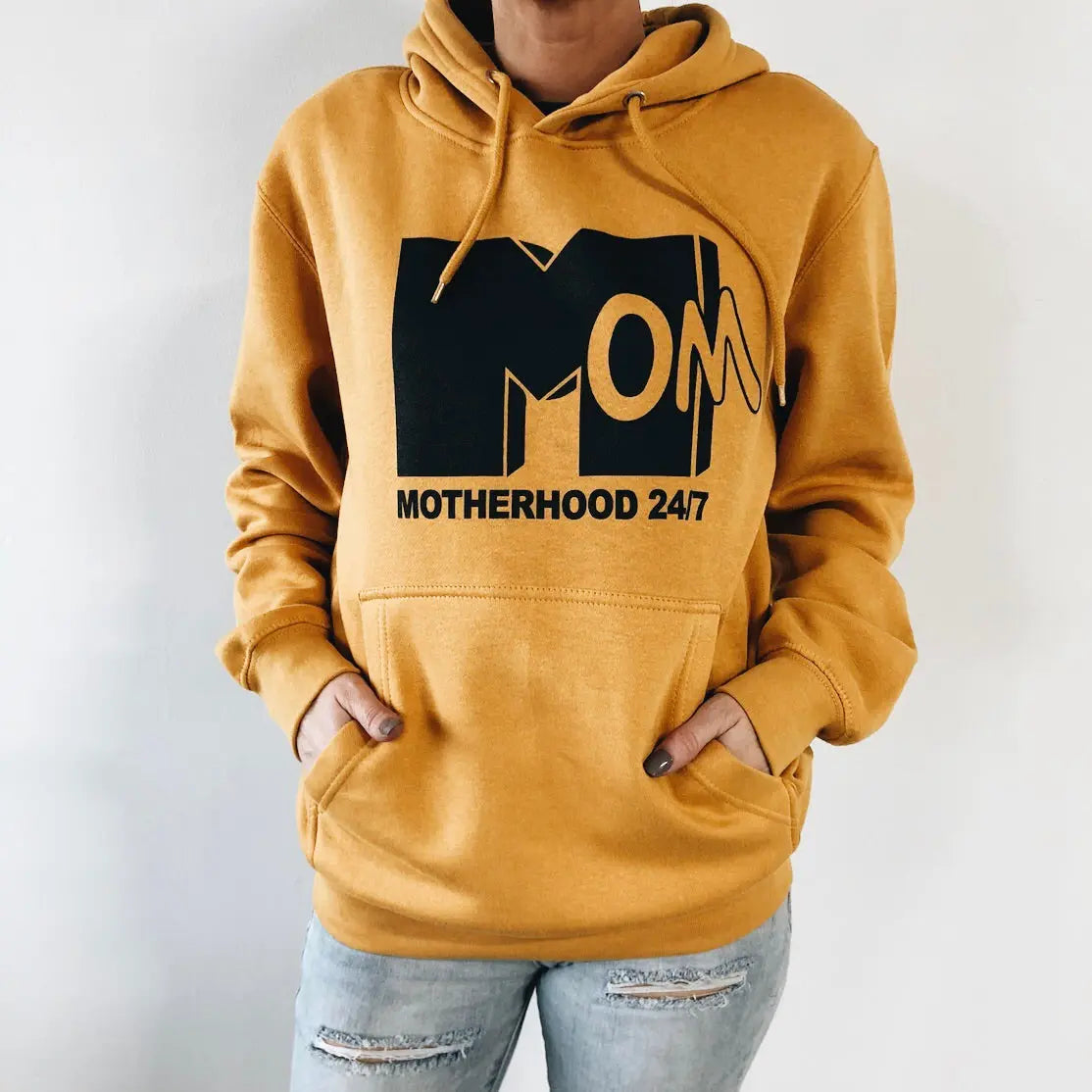 A stylish mom rocks the Motherhood 24/7 • Mustard Hoodie, fleece-lined with bold black MOM and Motherhood 24/7 lettering, hands in the front pocket, teamed effortlessly with light blue ripped jeans.