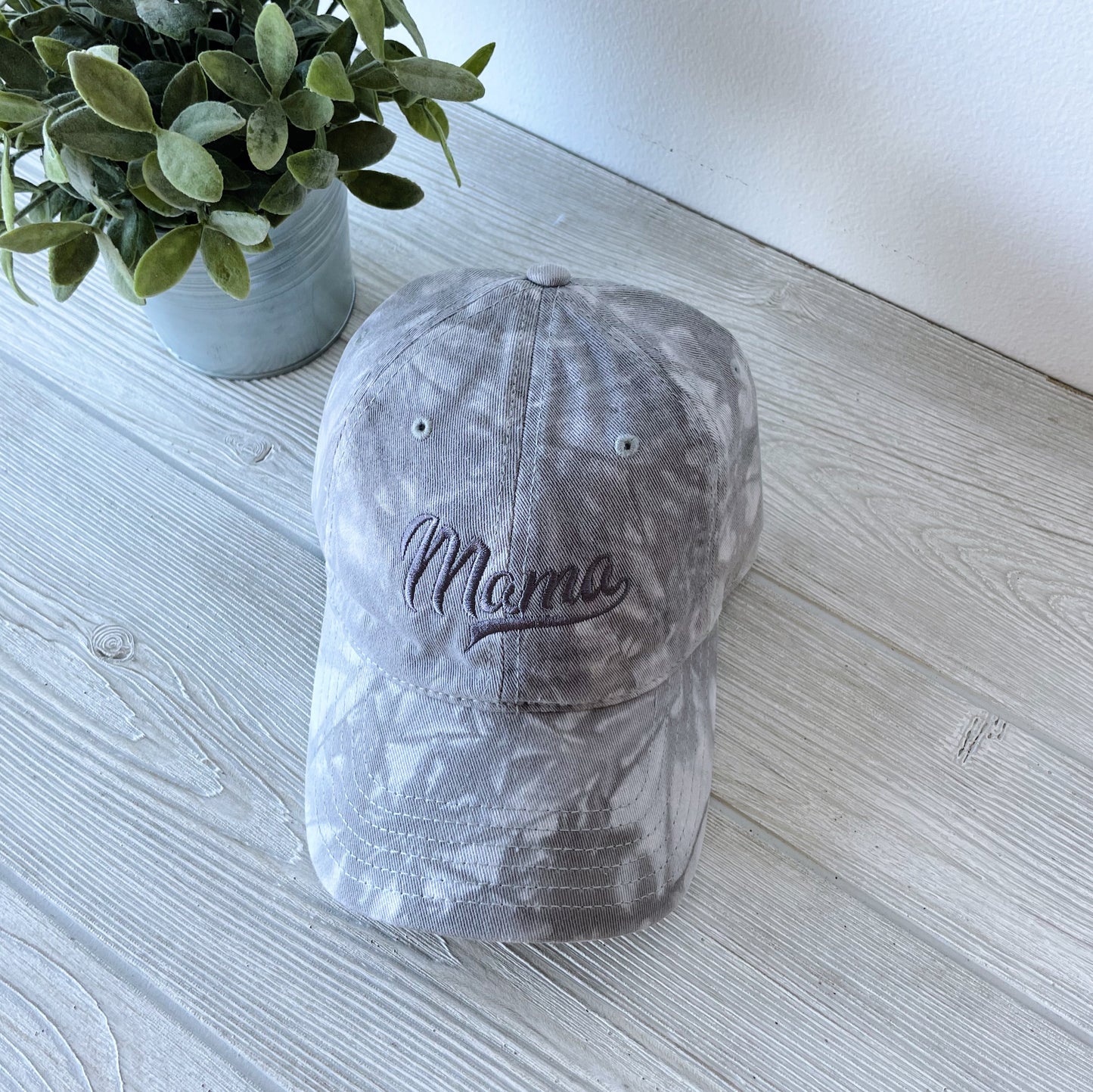 The Tonal Tie Dye Hat • Gray is a unisex twill baseball cap with Mama embroidered in script on the front, featuring a vintage-inspired gray tie-dye design for effortless charm. Shown on a light wooden surface beside a small potted plant.