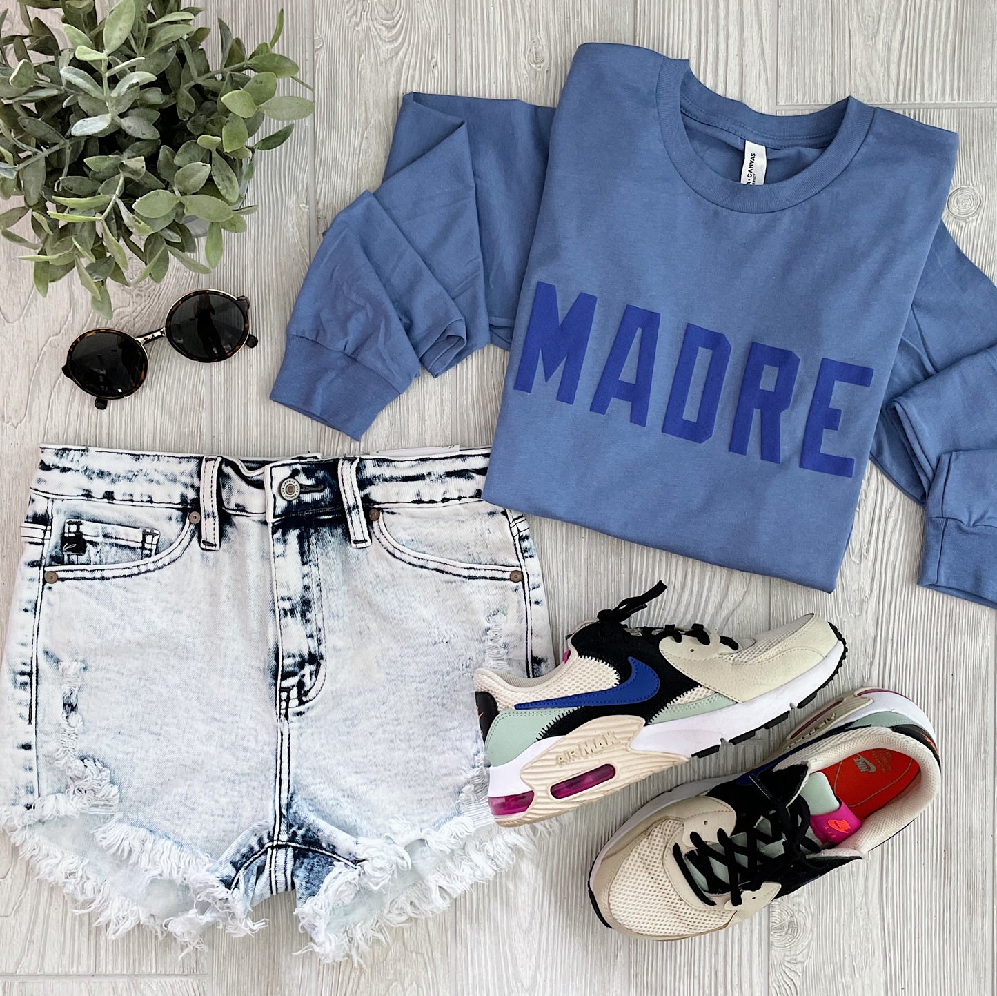 A flat lay of a casual outfit features the MADRE • Periwinkle Long Sleeve, light denim frayed shorts, white sneakers with colorful accents, black sunglasses, and a small green plant.