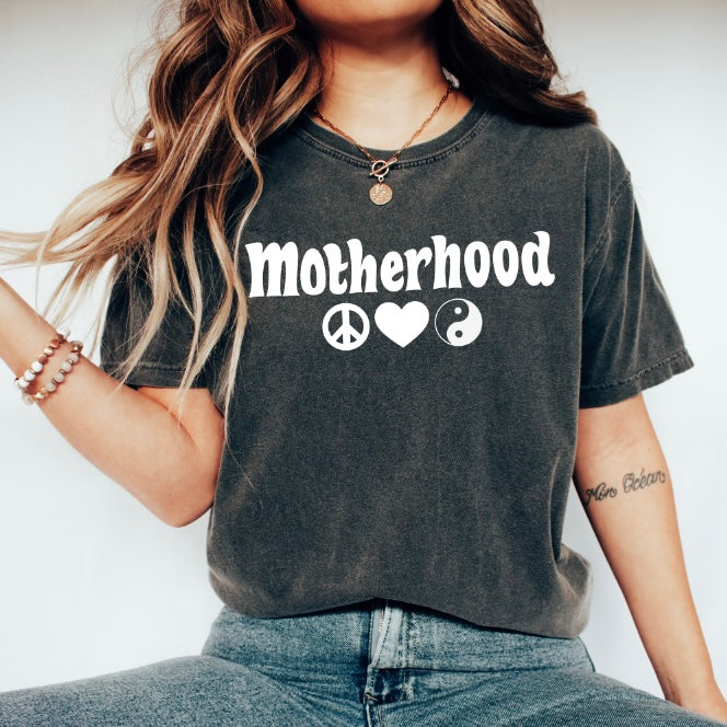 A woman with wavy hair, necklace, bracelets, and a small tattoo styles the Peace & Love • Comfort Colors Tee in dark gray, featuring Motherhood with peace, heart, and yin-yang symbols on soft ring-spun cotton.