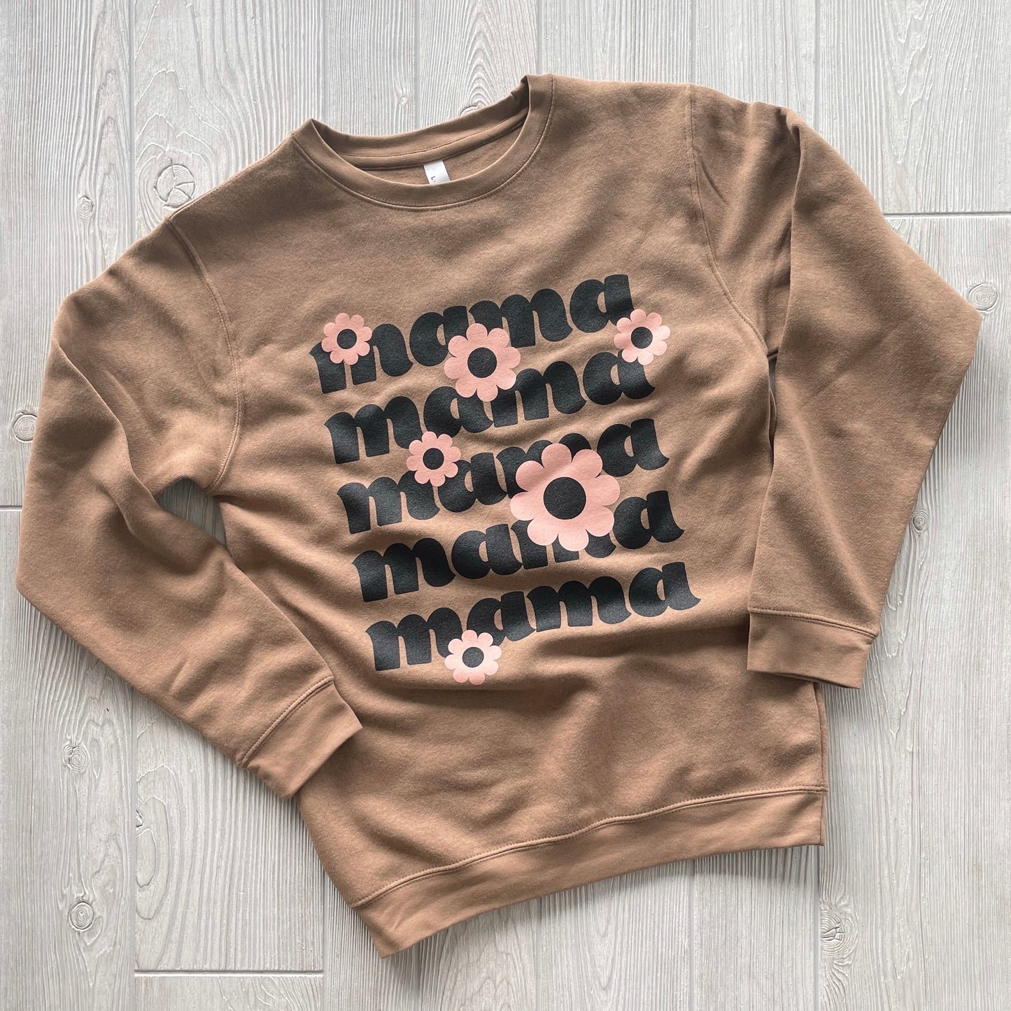 The Daisy Mama • Coyote Pullover is a brown sweatshirt featuring mama repeated five times in bold black letters, each line adorned with pink and beige flowers. Made of soft combed ringspun cotton and shown laid flat on a light wood floor.