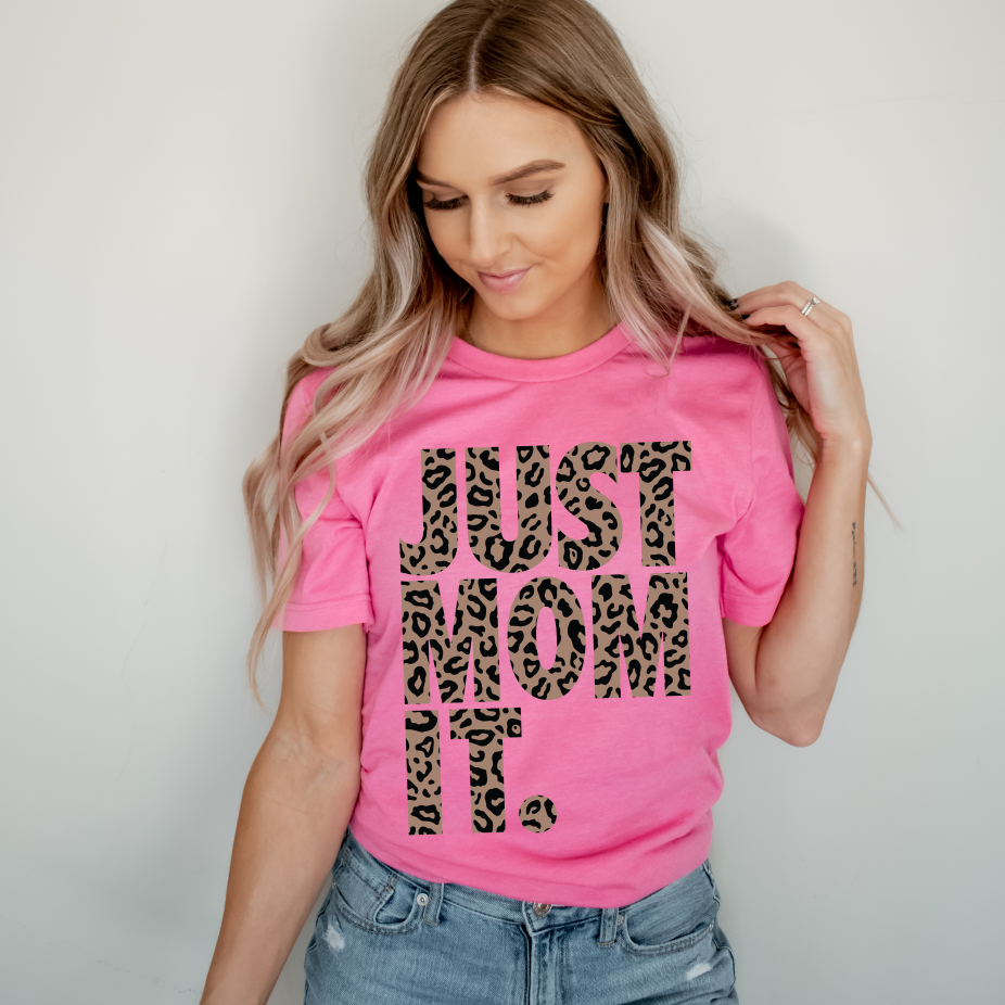A woman with long, wavy blonde hair wears the JUST MOM IT • Pink Tee Shirt in neon pink with bold cheetah print letters, paired with light blue jeans, standing against a plain light background.