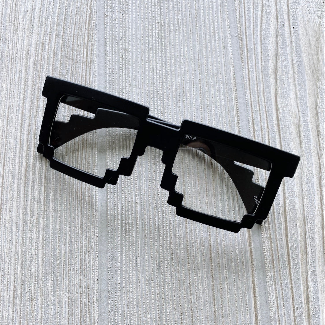 The Minecraft Glasses • Kids feature black, pixelated, blocky frames inspired by retro 8-bit video game graphics. Displayed on a light-textured wooden surface. Please note: this item is final sale.