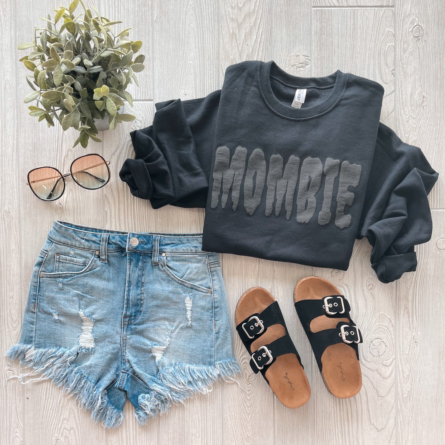 Flat lay of a casual outfit featuring the MOMBIE • Black Tonal Pullover, distressed denim shorts, black sandals with buckles, sunglasses, and a small potted plant on a light wooden surface.