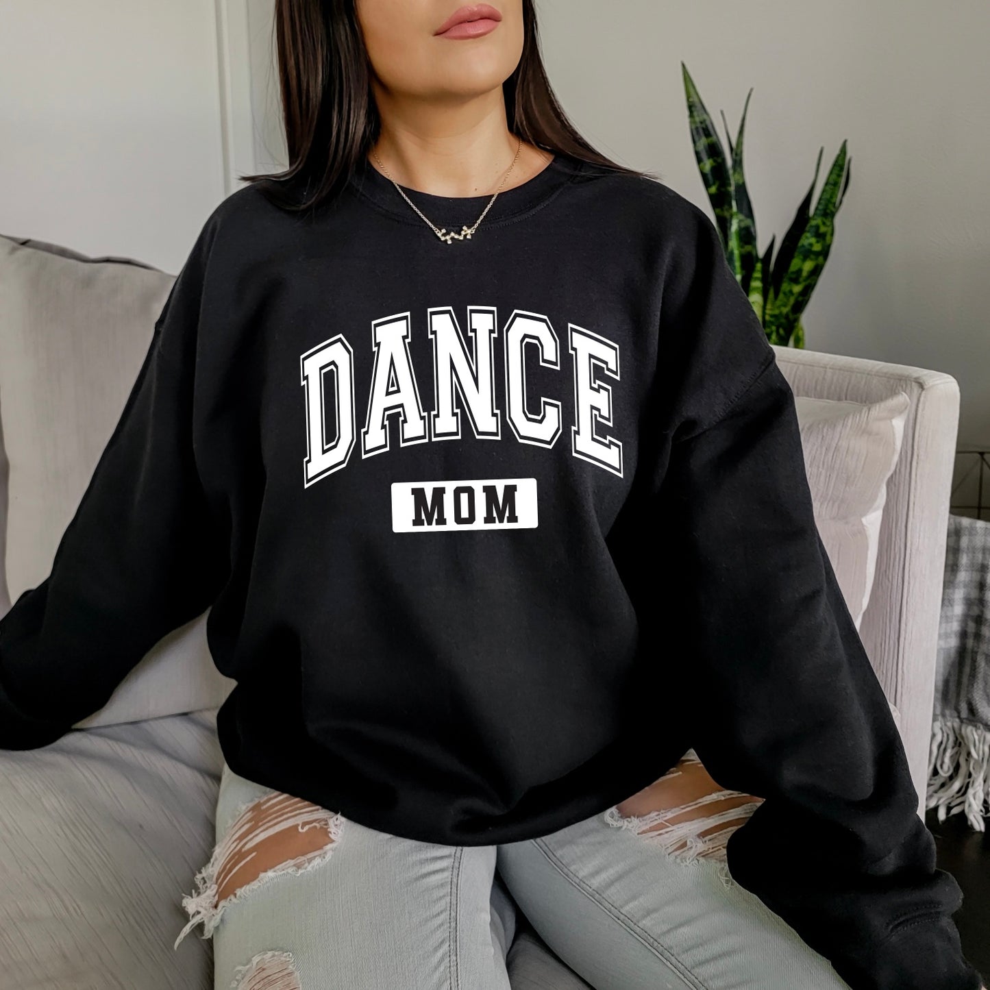 A woman relaxes on a light couch wearing the Dance Mom • Black Pullover from the Sports Mom Collection, featuring bold “DANCE MOM” lettering, paired with light ripped jeans. A green plant and gray blanket accent the background.