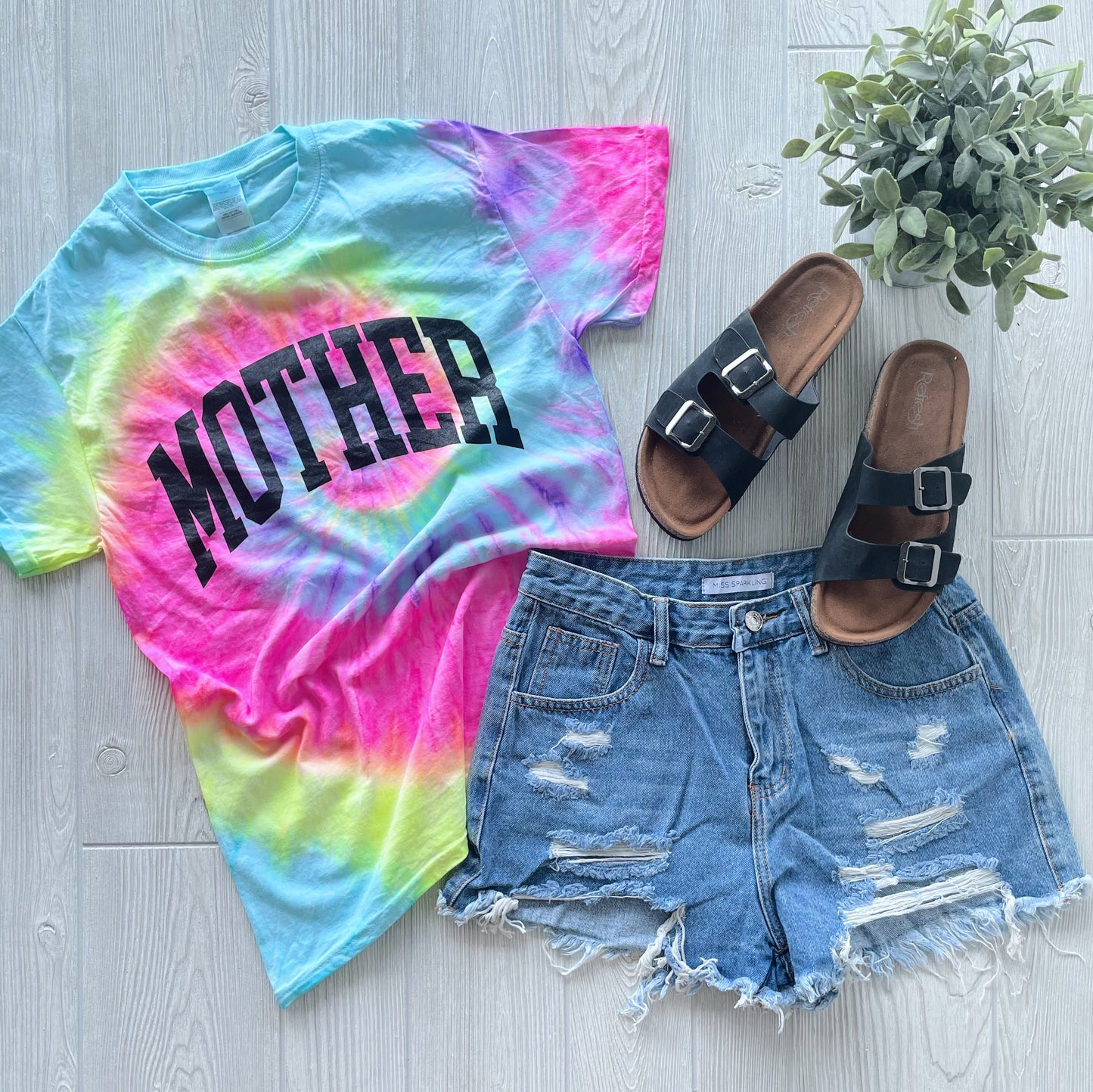 The MOTHER • Blast Tie Dye T-shirt, made from 100% cotton and featuring bold black MOTHER lettering, is displayed beside distressed denim shorts, black sandals, and a small green potted plant on a light wood floor.