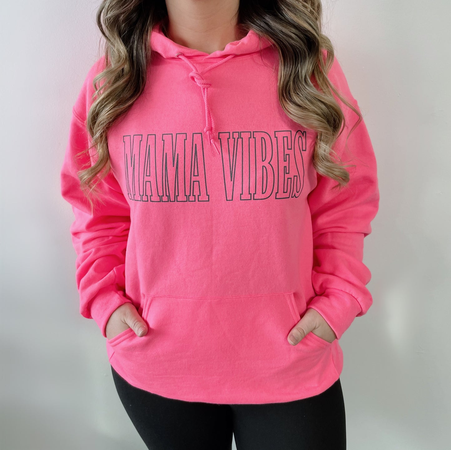 Someone wears the MAMA VIBES • Hot Pink Hoodie, featuring bold lettering and a comfy unisex fit, standing against a plain light background.