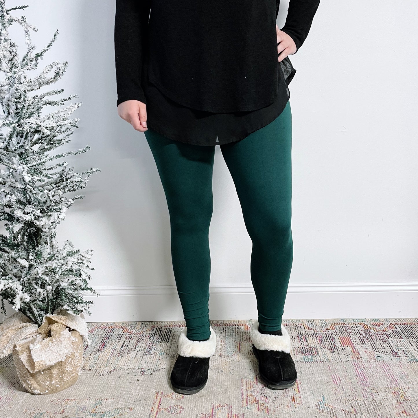 Wearing a plus size black brushed jersey top, black slippers with white fur trim, and Favorite Leggings in Forest Green, a person stands indoors by a small snow-dusted artificial Christmas tree in a burlap sack.