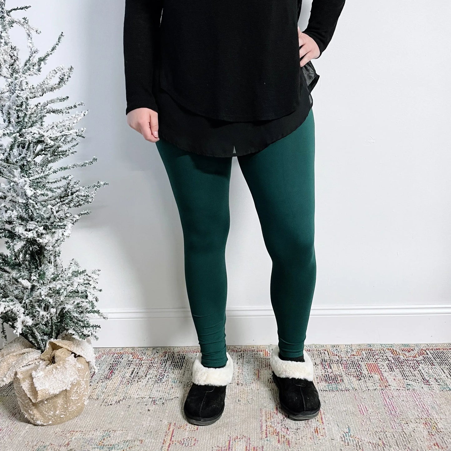 Wearing a plus size black brushed jersey top, black slippers with white fur trim, and Favorite Leggings in Forest Green, a person stands indoors by a small snow-dusted artificial Christmas tree in a burlap sack.