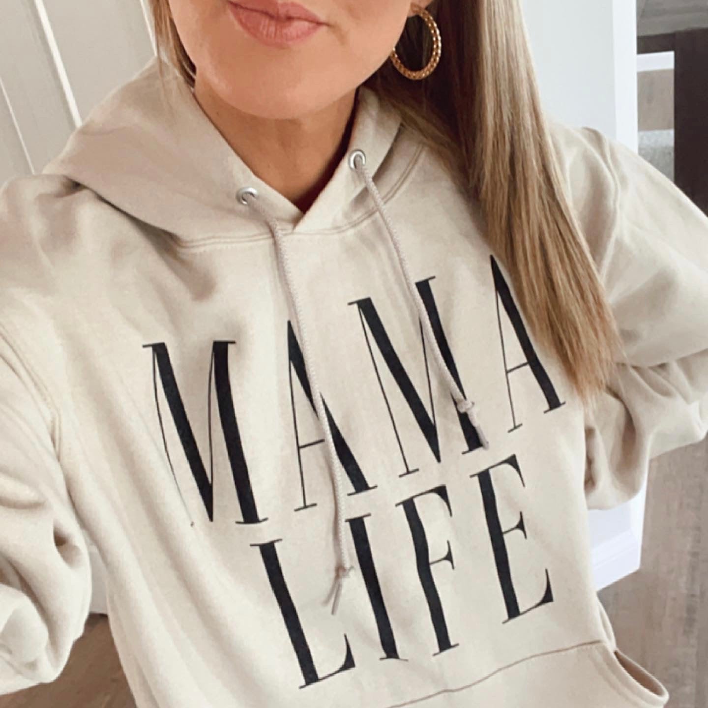 A woman models the Mama Life • Sand Hoodie, a cozy unisex fit from Mystery Hoodies. Only her lower face and torso are shown, highlighting the MAMA LIFE print on the front. She pairs it with straight light brown hair and hoop earrings.