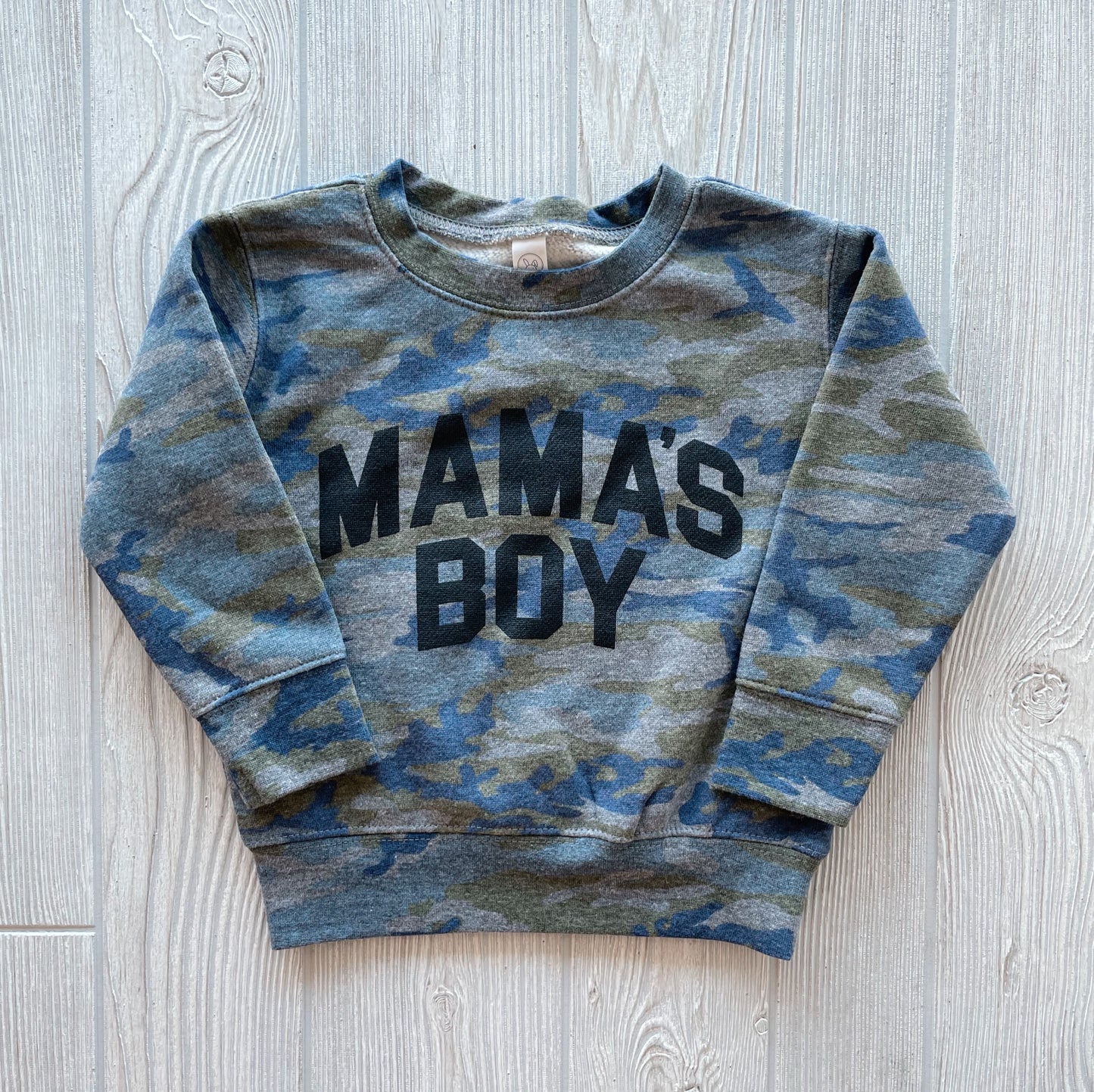 The MAMAS BOY • Camo Pullover is a unisex, long-sleeved toddler sweatshirt with a blue and green camo pattern and bold black MAMAS BOY text on the front, shown laid flat on a light wooden surface.