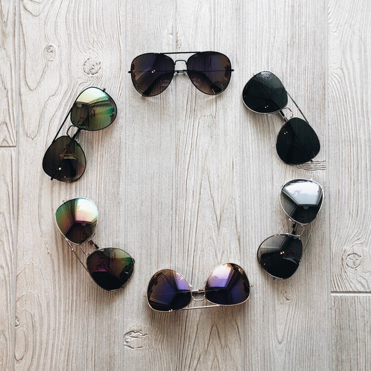 Six pairs of XL Avi Sunnies • Adult arranged in a circle on a light wooden floor, each pair facing outward to display different colorways.