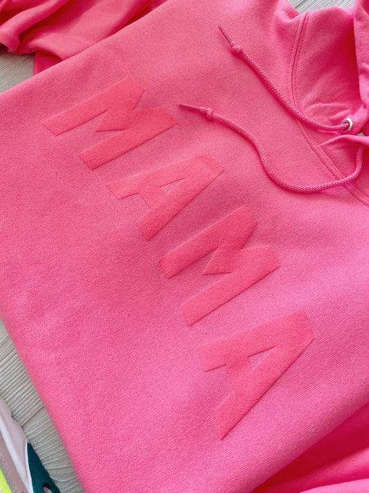 The MAMA arch • Neon Pink Tonal Hoodie features a unisex fit, suede “MAMA” arch print in bold raised letters across the chest, and visible drawstrings. Shown here on a light surface, it blends comfort with standout neon pink style.