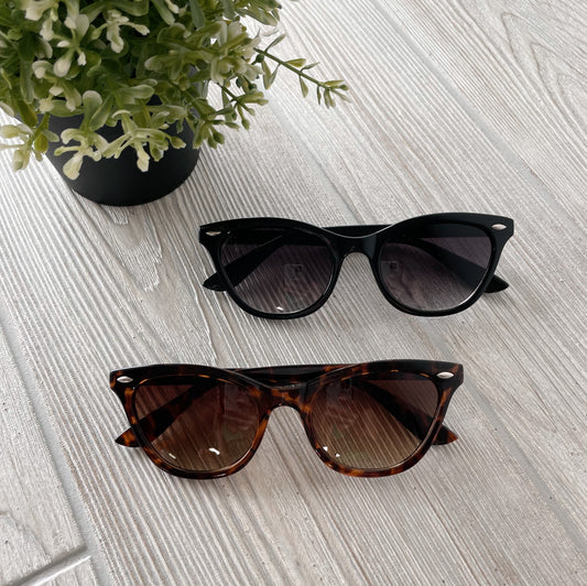 Two pairs of Bambi Sunglasses • Adult, one black and one tortoiseshell with UV400 protection, are displayed side by side on a light wooden surface next to a small potted green plant.