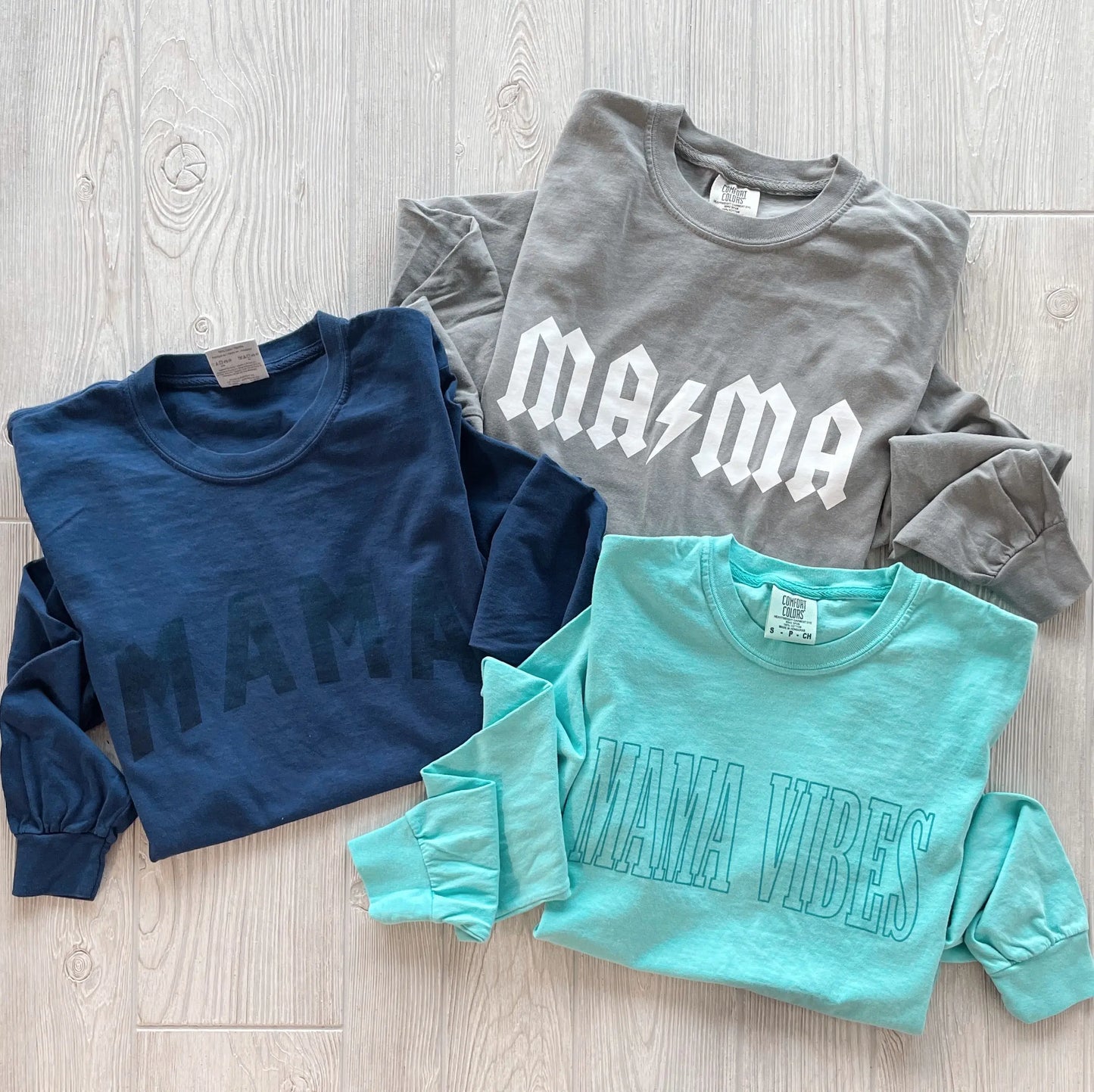 Three long sleeve tees on a light wood floor: the MAMA • Tonal Navy Long Sleeve, a gray shirt with “MAMA” in white and a lightning bolt, and a teal garment-dyed tee with “MAMA VIBES” in outlined letters.