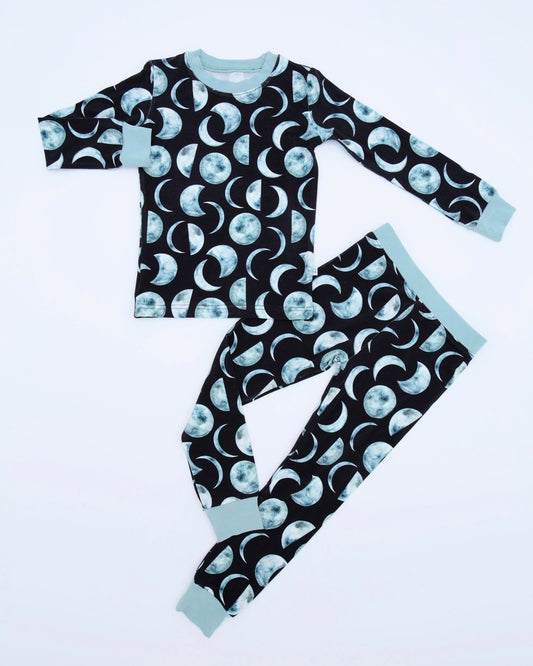 The Moon Child • 2 Piece Bamboo Set (Classic Fit) features a cozy long-sleeve design with a dark background, blue and white all-over moon phase pattern, and light blue cuffs on the sleeves, collar, and pant legs.