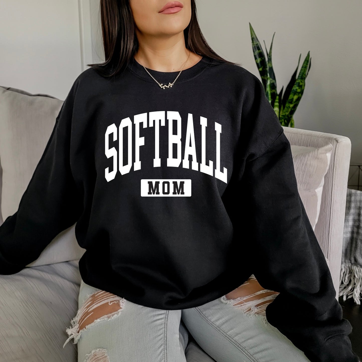 A woman sits on a couch wearing the Softball Mom • Black Pullover, a unisex fit sweatshirt with bold white SOFTBALL MOM lettering, paired with ripped light blue jeans. A green plant is seen in the background.